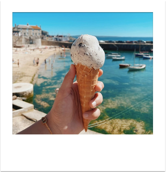 Holding an ice cream cone overlooking a sunny Cornish harbour with moored boats.